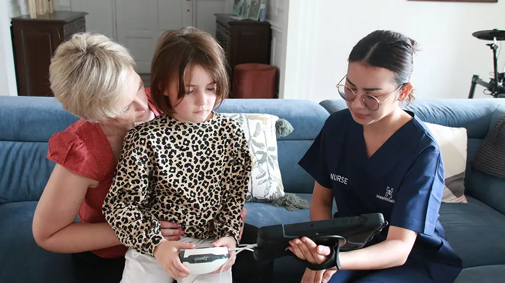 Healthcare nurse demonstrates medical device to grandmother and child on couch