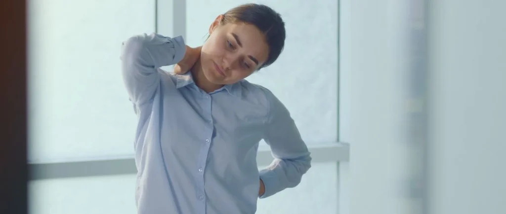 Woman in blue shirt stretching neck at office window, experiencing office syndrome pain